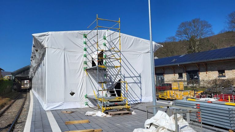 Weiß verhülltes Gebäude am Bahnsteig mit Gerüst, Gleis links und Solarmodulen.