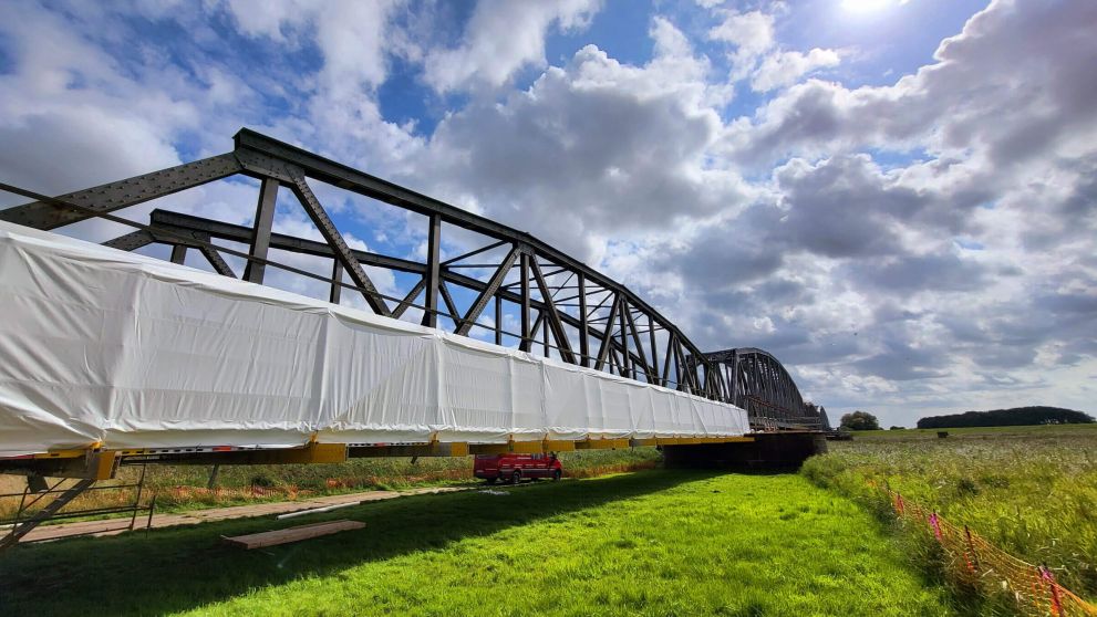 Stählerne Fachwerkbrücke mit teilweise weiß verhüllter Fahrbahn über grüner Wiese.