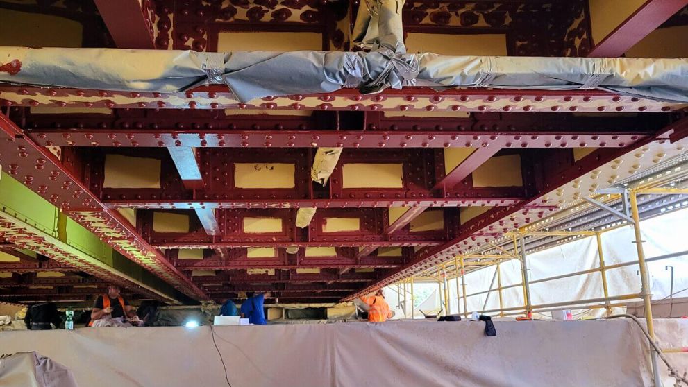Unter einer rot gestrichenen Stahlträgerkonstruktion mit Nieten stehen Gerüste und arbeiten Handwerker.