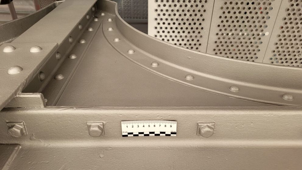 Grau lackierter Metallträger mit Nieten, drei großen Sechskantmuttern und kleinem Maßstabsaufkleber.