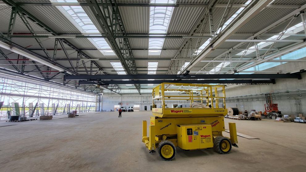 In der großen hellen Industriehalle steht eine gelbe Scherenarbeitsbühne, in der Ferne eine Person.
