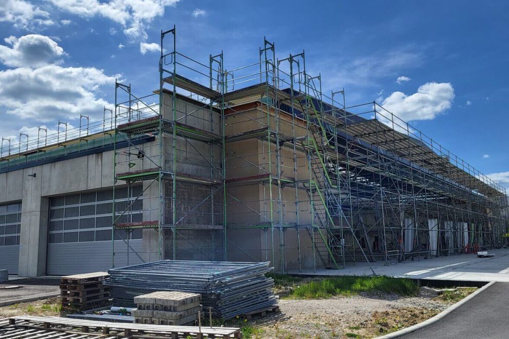 Große Halle mit eingerüsteter Fassade, mehreren geschlossenen Rolltoren und Materialstapeln.