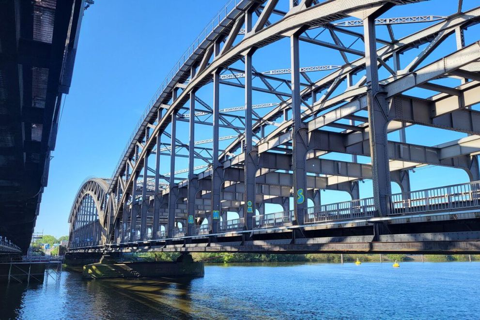 Große stählerne Bogenbrücke mit offenem Fachwerk über ruhigem Fluss, linker Brückenrand sichtbar.