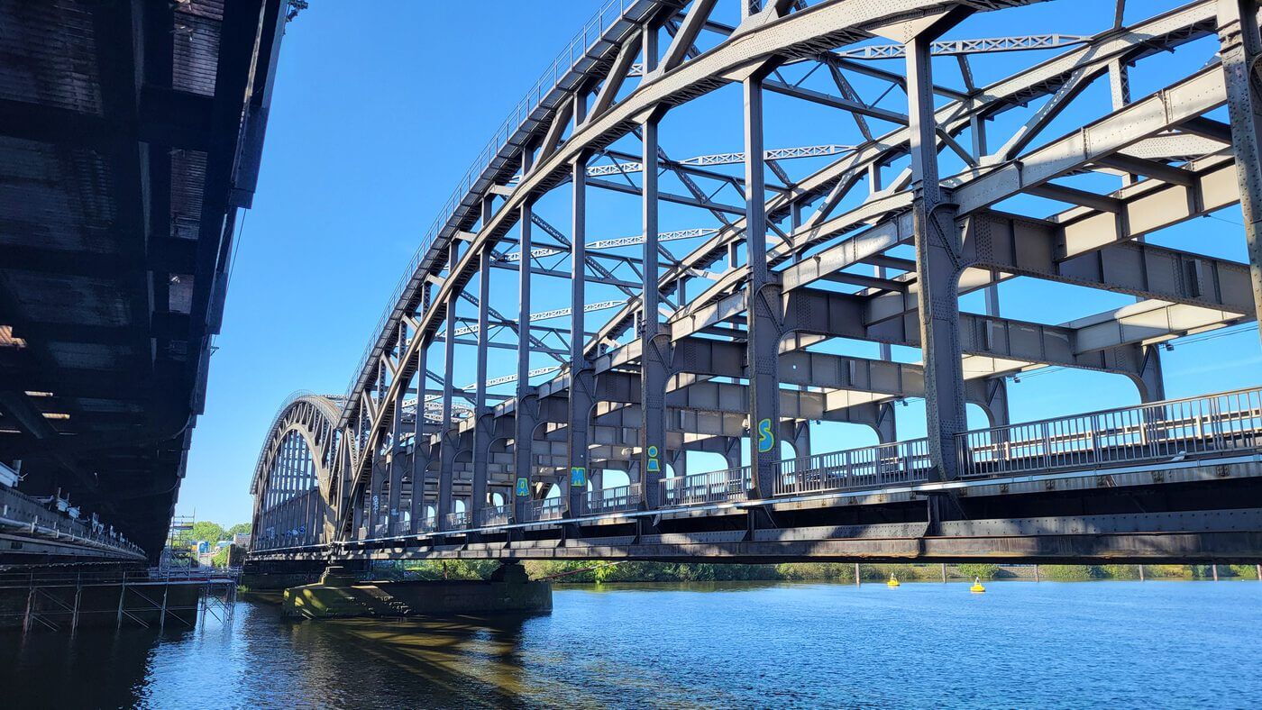 Große stählerne Bogenbrücke mit offenem Fachwerk über ruhigem Fluss, linker Brückenrand sichtbar.